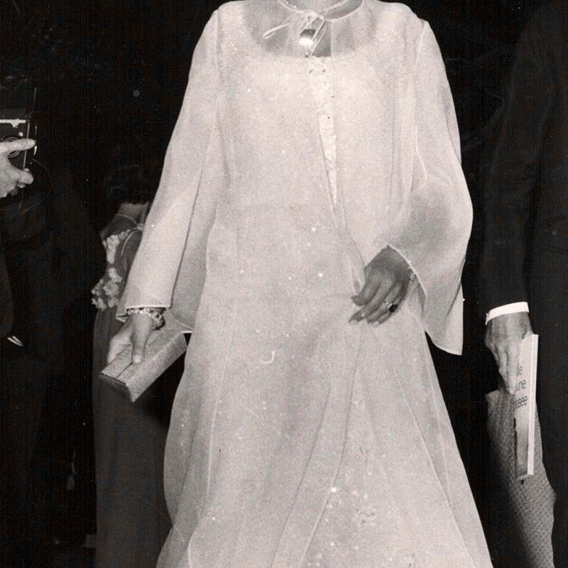 Frau in eleganter, weißer Abendgarderobe mit langen Ärmeln, umgeben von Menschen und Kameras, 1960er Jahre.