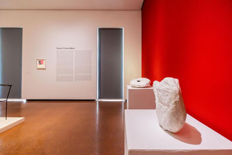 Ausstellungsraum mit roter Wand, Skulpturen auf Podesten und einer Wandtafel zu Franz West.