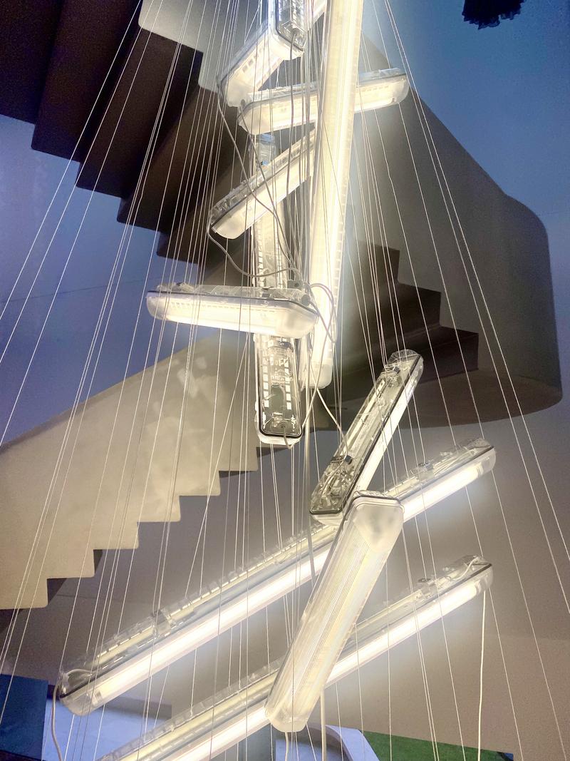 Hängelampe mit mehreren leuchtenden Röhren, die über eine Treppe in einem modernen Raum angeordnet sind.