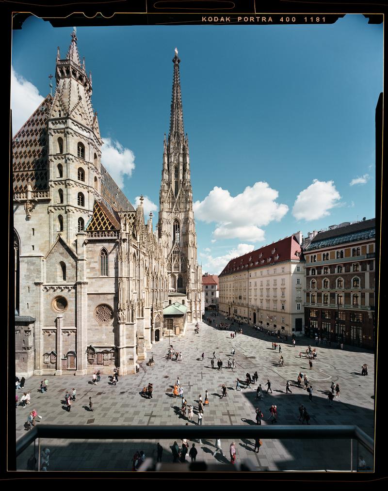 Auf dem Foto sehen wir den Stephansdom in Wien.
Das Bild wurde von einem Balkon aus gemacht.

Der Stephansdom ist eine sehr alte Kirche.
In der Mitte sieht man einen sehr hohen, spitzen Turm.
Das Dach hat bunte Ziegel. Es sieht schön und besonders aus.
Links ist die Kirche, rechts sind andere Häuser.
Auf dem Platz davor sind viele Menschen.

Der Himmel ist blau. Es gibt ein paar weiße Wolken.

Das Foto wurde mit einer alten Kamera gemacht, die einen Film benutzt.

Der Fotograf heißt Stefan Oláh.
Die Farben im Bild sind warm und angenehm.