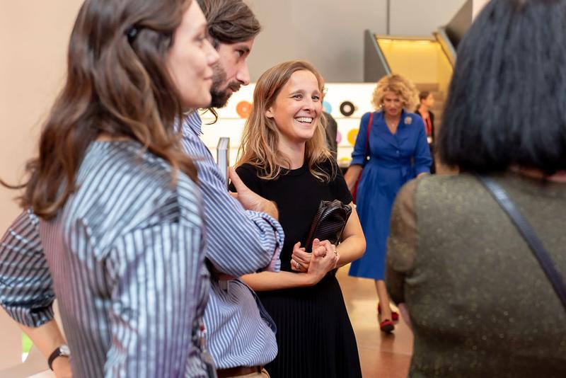 Auf dem Bild sieht man eine Gruppe von Menschen in der Heidi Horten Collection.
Alle schauen sich etwas an und unterhalten sich.

In der Mitte des Fotos steht eine Frau mit blondem Haar.
Sie trägt ein schwarzes Kleid und lächelt freundlich.
Um sie herum stehen mehrere Personen – sie sehen interessiert und entspannt aus.

Im Hintergrund sieht man eine Frau im blauen Kleid, die ebenfalls durch den Raum geht.
Die Stimmung auf dem Foto wirkt offen, freundlich und neugierig