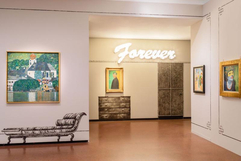 Auf dem Foto sieht man einen Teil der Ausstellung "Klimt⇄Warhol" im Museum der Heidi Horten Collection in Wien.

Links, hängt ein Gemälde von Gustav Klimt.
Darauf sieht man den Attersee, einen See in Österreich.
Das Bild zeigt Wasser, Häuser und Natur in hellen Farben.

Die Verzierungen an den Wänden und die gemalten Möbelstücke neben und unter den Kunstwerken hat ebenfalls Markus Schinwald gestaltet.

Hinten an der Wand, über einem Bild, leuchtet in großen weißen Buchstaben das Wort „Forever“.
Diese Leuchtschrift ist ein Kunstwerk von Tim Noble & Sue Webster.
Darunter hängt ein Selbstporträt von Edvard Munch.