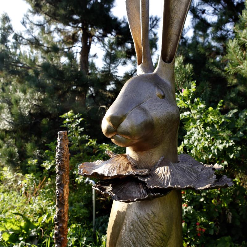 Bronzeskulptur eines Kaninchens mit großen Ohren, einem Kragen aus Blättern und einem Stock in der Hand, umgeben von Pflanzen.