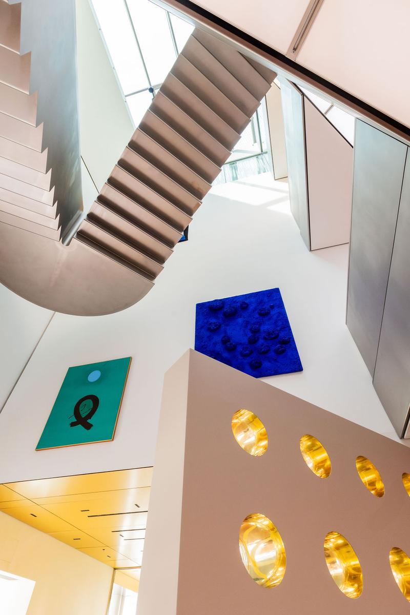 Auf dem Foto sieht man einen Blick nach oben im Museum der Heidi Horten Collection in Wien.
Man erkennt eine moderne Treppe aus Metall und große, helle Wände.

Links an der Wand hängt ein grünes Bild mit einem schwarzen Zeichen und einem blauen Kreis.
Dieses Kunstwerk heißt „Blue Orb“ und ist von Adolph Gottlieb.

Daneben, weiter oben, sieht man ein kräftig blaues Kunstwerk mit Schwämmen darauf.
Das ist das allererste Schwammrelief von Yves Klein.
Er hat echte Schwämme mit tiefblauer Farbe eingefärbt und auf die Leinwand geklebt.
Das Werk ist sehr besonders, weil Yves Klein für diese intensive blaue Farbe berühmt wurde.

Unten im Bild sieht man einen Teil der Bullaugenwand von Markus Schinwald.
Die runden, gold glänzenden Fenster gehören zu einem Gestaltungselement.