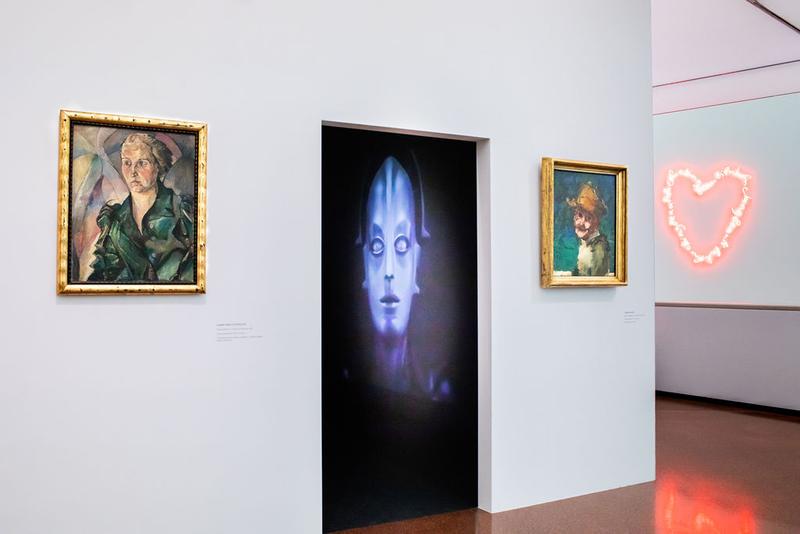 Auf dem Foto sieht man einen Durchgang in der Ausstellung „Experiment Expressionismus. Schiele meets Nosferatu“.
Im Raum hinter dem Durchgang sieht man ein großes, leuchtendes Bild vom Maschinenmenschen aus dem Film „Metropolis“ (1927).

Links und rechts vom Durchgang hängen zwei Porträts in goldenen Rahmen:
Links ist ein eckiges und ernstes Porträt einer Frau.
Rechts ist ein kleineres, lockerer gemaltes Porträt mit blaugrünem Hintergrund und gelbem Hut – es ist das Selbstporträt von Herbert Boeckl.

Rechts im Hintergrund sieht man ein leuchtendes Herz aus rosa Neonlicht an der Wand.