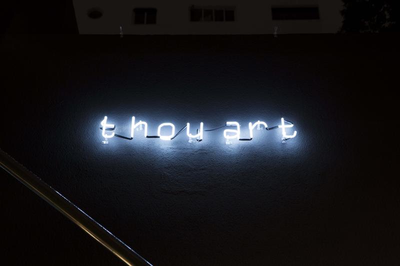 Neonlichtschriftzug "thou art" auf einer dunklen Wand, mit sanfter Beleuchtung und minimalistischem Design.