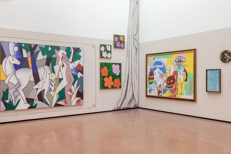 Auf dem Foto sieht man einen Teil der Ausstellung "Klimt⇄Warhol" im Museum der Heidi Horten Collection in Wien.

Die Aufnahme zeigt eine Ecke des Ausstellungsraums,
an der zwei Wände aufeinandertreffen. An diesen Wänden hängen mehrere bunte Gemälde.

Ganz links sieht man ein großes Bild von Roy Lichtenstein.
Es zeigt mehrere weiße Rehe in einer abstrakten Landschaft,
mit starken Farben und schwarzen Umrisslinien – typisch für Lichtensteins Stil, der an Comics erinnert.

Rechts daneben, übereinander gehängt, sieht man drei kleine Bilder mit bunten Blumen.
Diese Blumendrucke stammen von Andy Warhol. Sie sind sehr farbenfroh, mit einfachen Formen und starkem Kontrast.

In der rechten Ecke hängt ein großes, gemeinsames Gemälde von Jean-Michel Basquiat und Andy Warhol.
Das Bild ist wild, bunt und voller Energie.
Man erkennt Figuren, Schrift und Gesichter.

Rechts daneben sieht man einen weiteren Druck von Andy Warhol –
ein kleines, hellblaues Bild, auf dem man Elisabeth Taylor als Kleopatra sieht.

Darüber, rund in gold ist ein weiteres Werk von Warhol:
ein Porträt von Jackie Kennedy, der früheren First Lady der USA.

Ein aufgemalter, grauer Vorhang trennt die beiden Wände optisch und erinnert an ein Theater oder eine Bühne.
Auch dieser Teil der Wandgestaltung gehört zum inszenierten Ausstellungsdesign von Markus Schinwald.