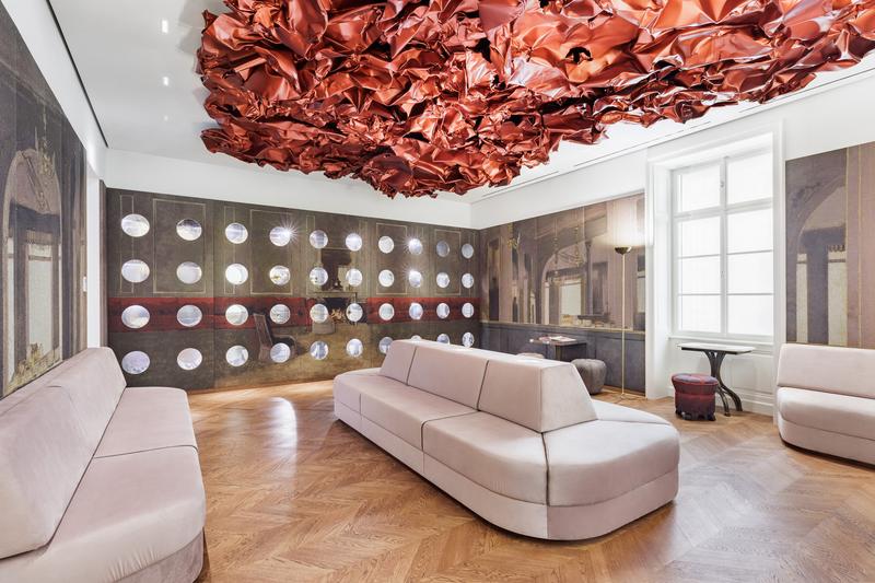 Auf dem Foto sehen wir den Tea Room im Museum der Heidi Horten Collection in Wien.

An der Decke hängt ein großes, rotes Kunstwerk von Hans Kupelwieser.
Es sieht aus wie zerknittertes Papier oder glänzender Stoff,
wurde aber aus Aluminium gemacht.

An der hinteren Wand gibt es viele runde Fenster – sie sehen aus wie Bullaugen.
Dahinter befinden sind kleine, kostbare Objekte aus der Sammlung von Heidi Horten.

Diese Vitrinenwand wurde von Markus Schinwald entworfen.
Auch die rosa Sitzmöbel in der Mitte des Raums gehören zu seinem Konzept.
Sie laden ein, sich hinzusetzen, zu schauen und zu entspannen.

Die Wände sind mit großen Stoffbildern bedeckt,
die an ein altes Café oder Schlosszimmer erinnern.